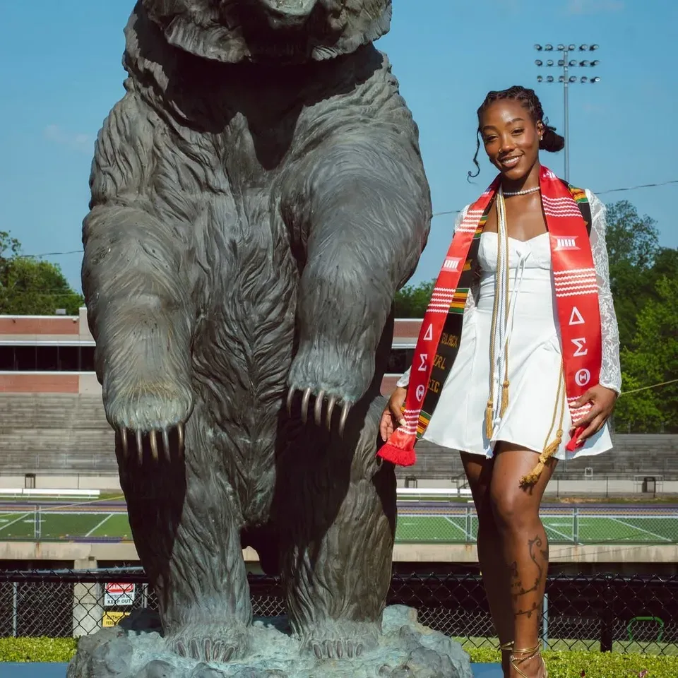 Hannah Smith in a white dress and graduation stole beside a bear statue at Miles College